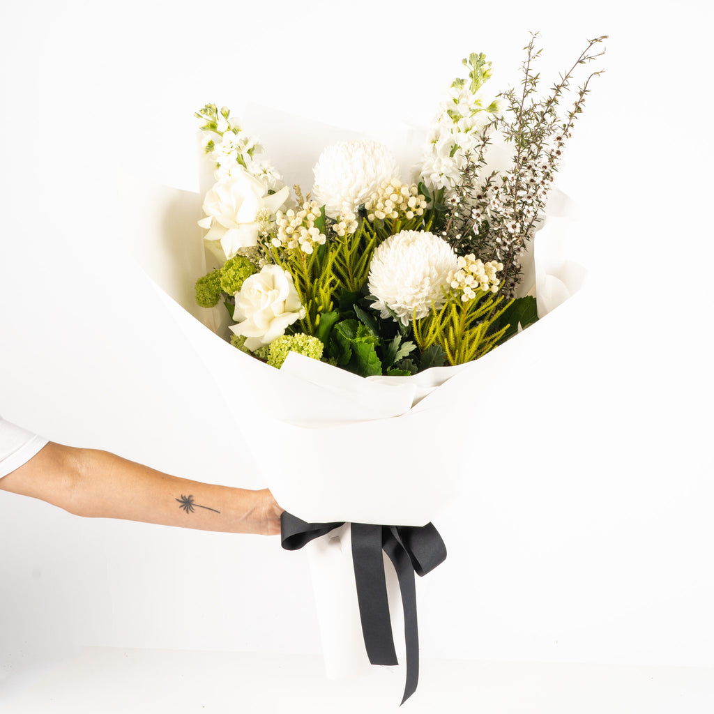 Fresh Whites n Greens Flower Subscription by Wild Poppies featuring a hand-tied bouquet of white and green blooms wrapped in white paper with a black ribbon.