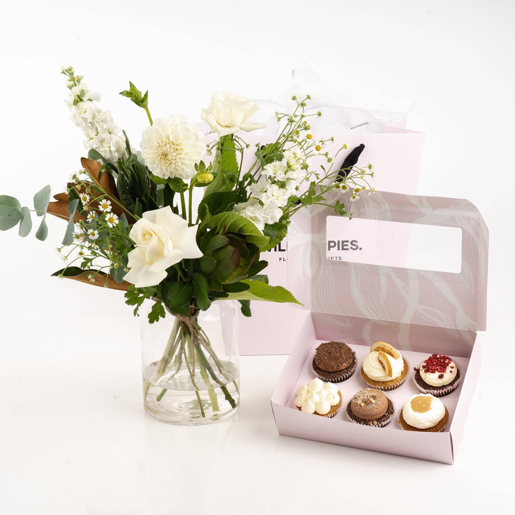 White n Greens with Gourmet Cupcakes gift set featuring a fresh white floral bouquet in a glass vase and six assorted handcrafted cupcakes in a pink box.