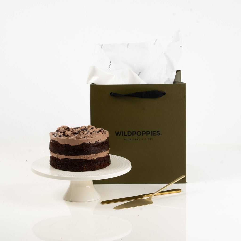 Chocolate cake on a white stand next to a green 'Wildpoppies' gift bag on a white background