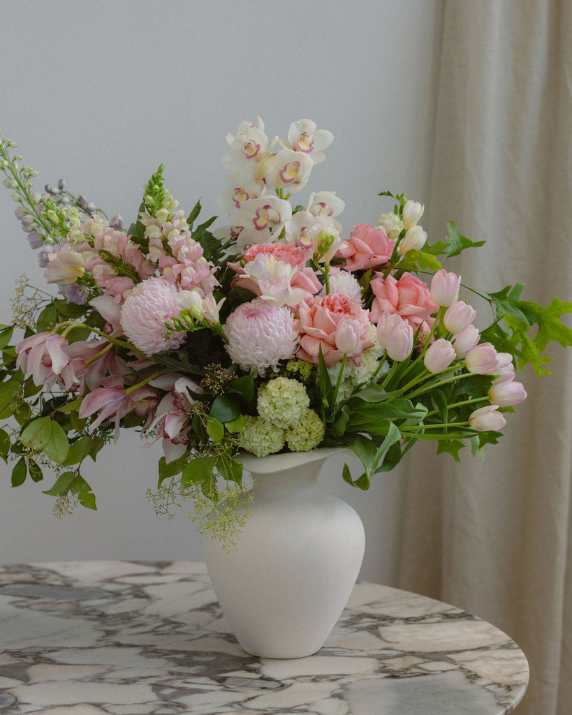 Bouquet of pink and white flowers in a white vase on a marble table with a beige curtain in the background.