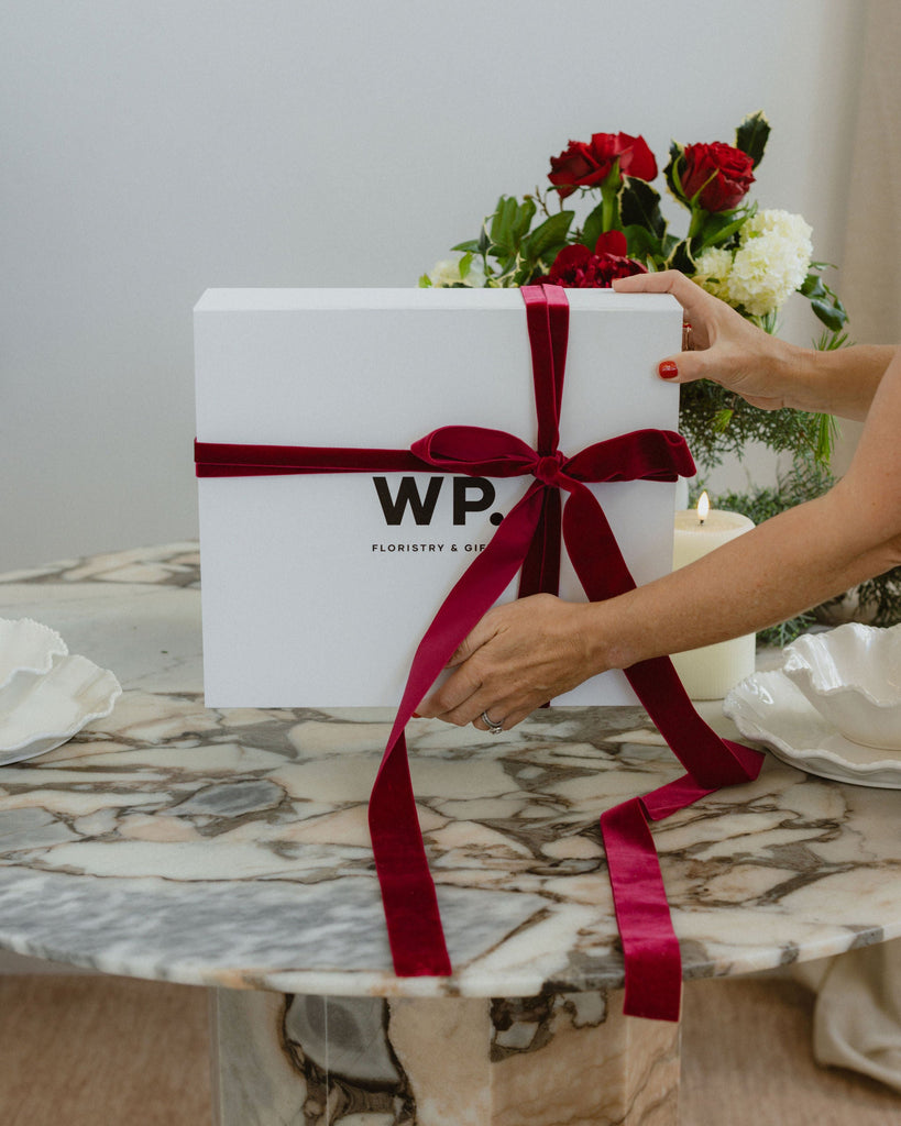 White gift box with red ribbon held by a person on a marble table with flowers in the background