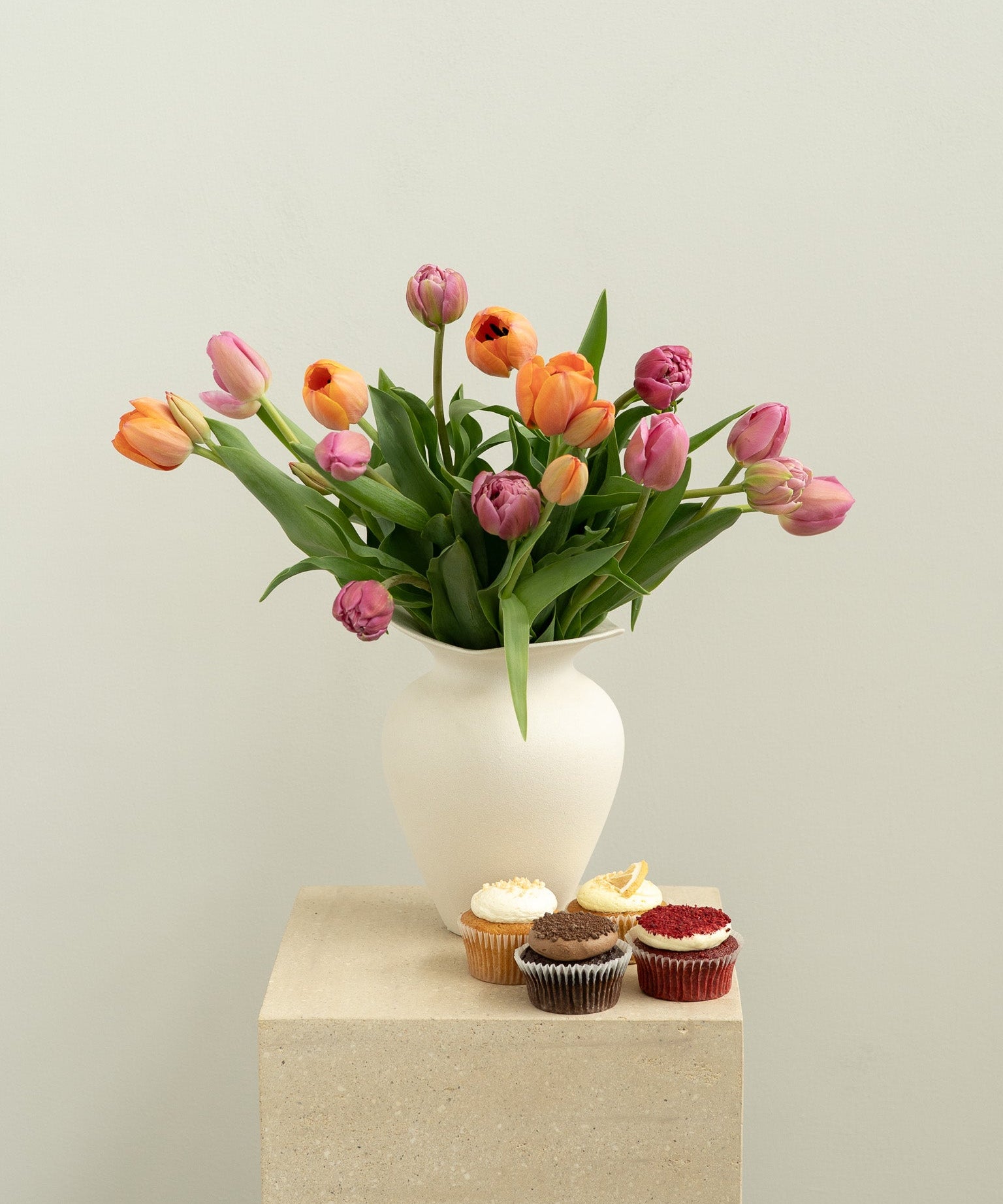 Vase of tulips with cupcakes on a pedestal against a plain background