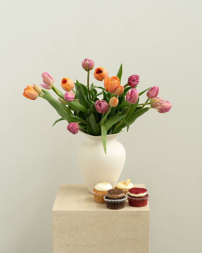 Vase of tulips with cupcakes on a pedestal against a plain background